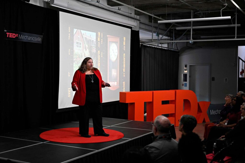 Woman in Red standing on stage doing a presentation - TEDx Mechanicsville sign in background