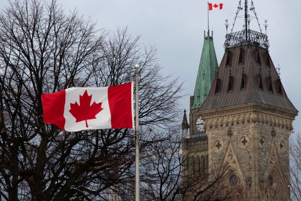 Canadian flag flying with the roof of parliament hill in the background.